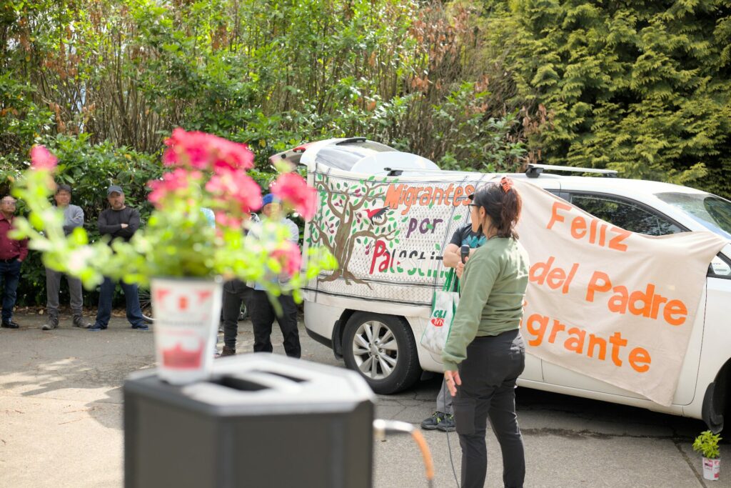 Un miembre de Fuerza Migrante platica con trabajadores agricolas migrantes durante el Dia del Padre Migrante 2023. En el fondo, dos mantas leen "Feliz Dia del Padre Migrante" y "Migrantes por Palestina".